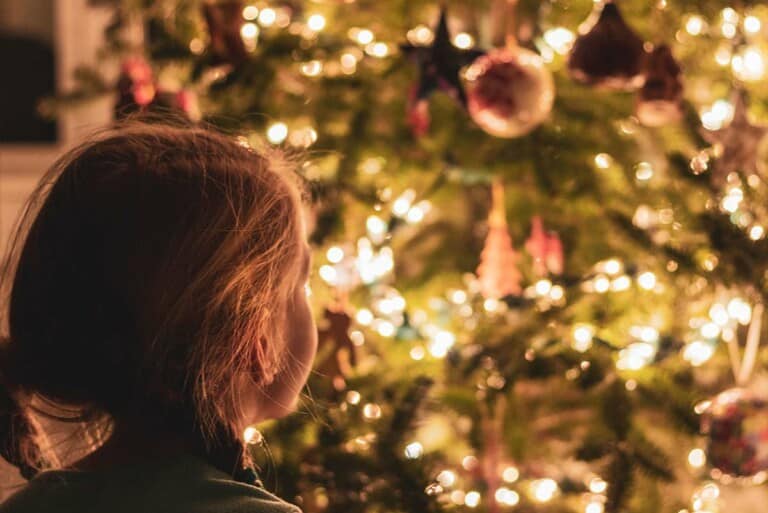 Ein Kind steht vor einem geschmückten Weihnachtsbaum mit Kugeln und Lichterketten.