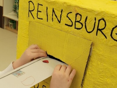 Ein Kind steckt einen Brief in einen selbstgebastelten, gelben Postbriefkasten.