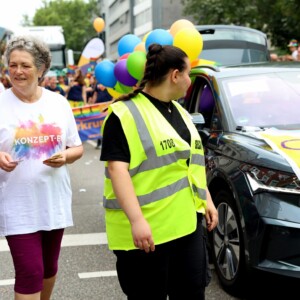 Frau Weegmann verteilt Brause beim CSD 24. Rechts davon läuft eine Ordnerin neben unserem Auto her.