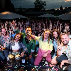 Die Band lächelt in die Kamera und im Hintergrund strecken die Besucher:innen die Hände in die Luft