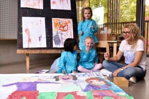 Eine Erzieherin mit blonden Haaren und Brille sitzt auf dem Boden und hat mit Kindern Spaß beim Malen.